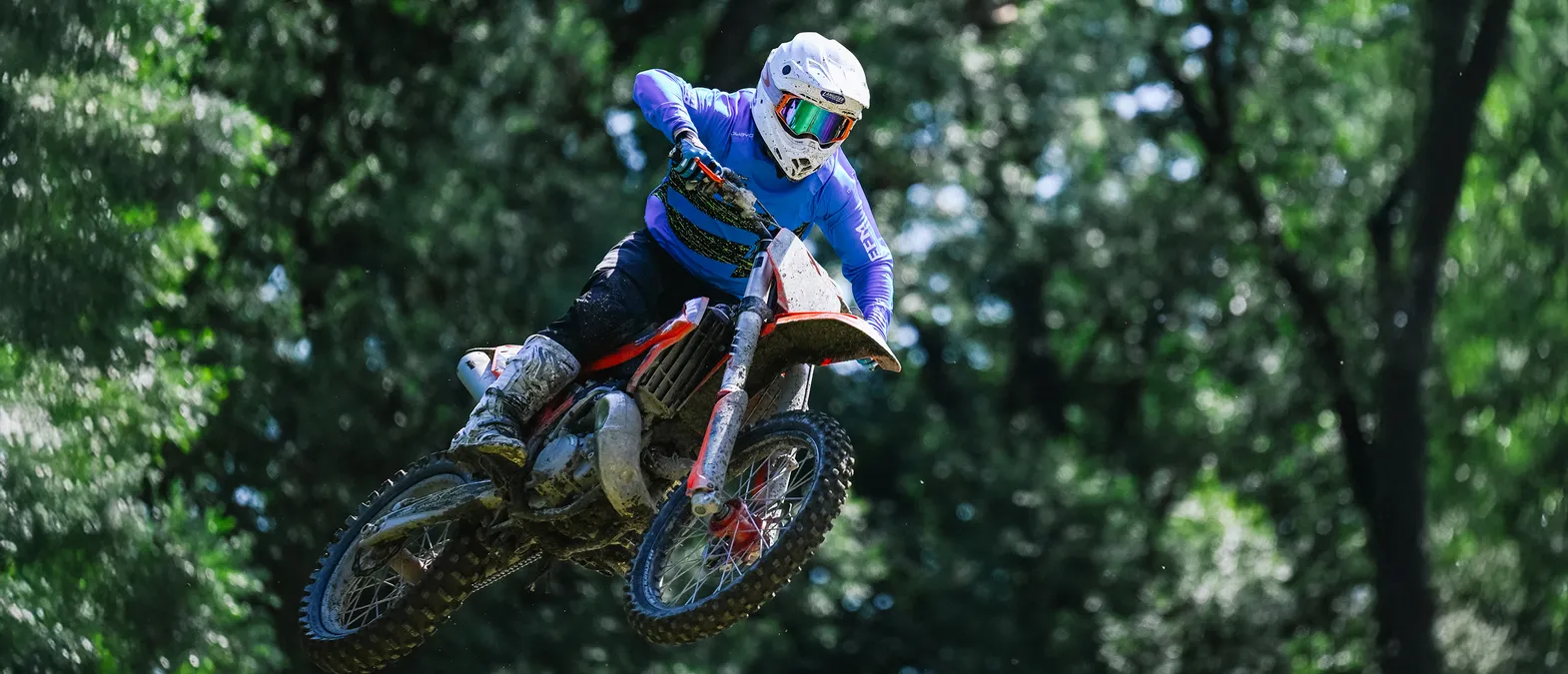 Un motocycliste en combinaison de course bleue à manches longues et casque blanc saute sur une moto dans une forêt.