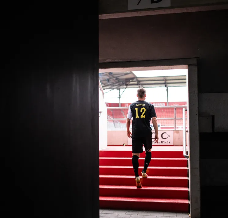 Un joueur de football en maillot noir avec l'inscription 'MIEYER 12' en jaune monte les marches rouges d'un stade.