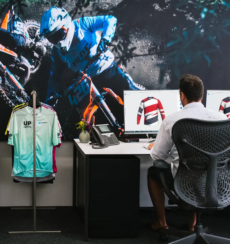 Un homme est assis à un bureau en train de concevoir un maillot de sport personnalisé sur un ordinateur, avec une grande image d'un motocycliste et un portant de vêtements de sport en arrière-plan.