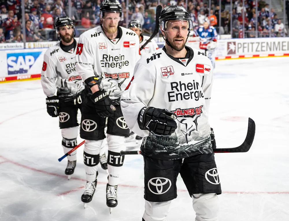 Trois joueurs de hockey sur glace en maillots blancs avec des logos noirs et des designs graphiques se tiennent sur la glace.