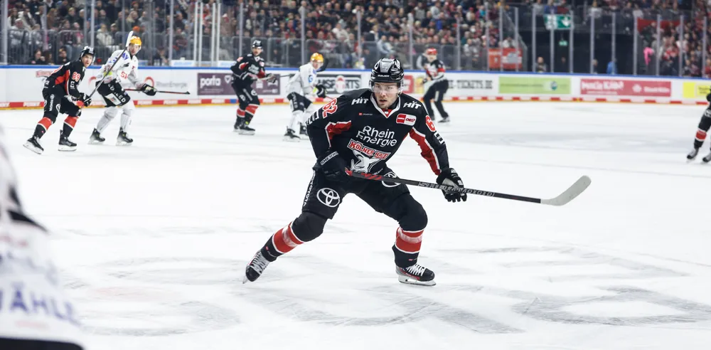 Un joueur de hockey sur glace portant un maillot noir avec des accents rouges et blancs, patinant sur la glace avec une crosse, tandis que des spectateurs acclament en arrière-plan.
