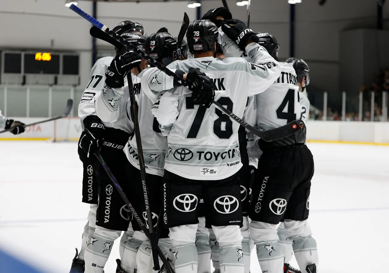 Joueurs de hockey sur glace en maillots blancs avec des numéros noirs et des logos de sponsors, se serrant dans les bras sur la glace.