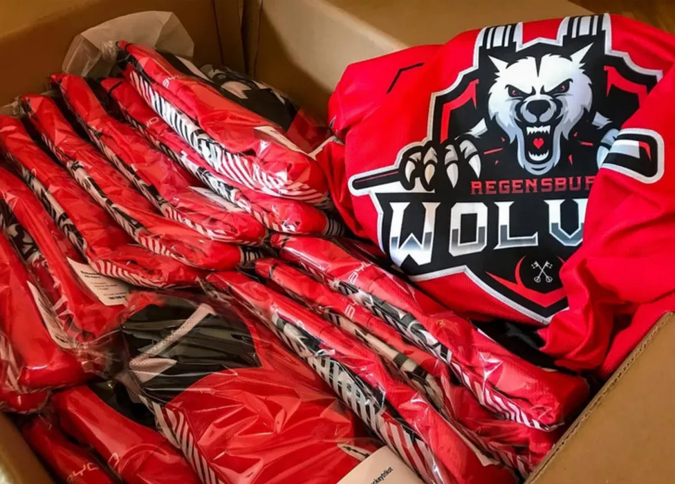 Une boîte remplie de maillots de sport rouges personnalisés, dont un avec un logo de loup et l'inscription 'Regensburg Wolves'.