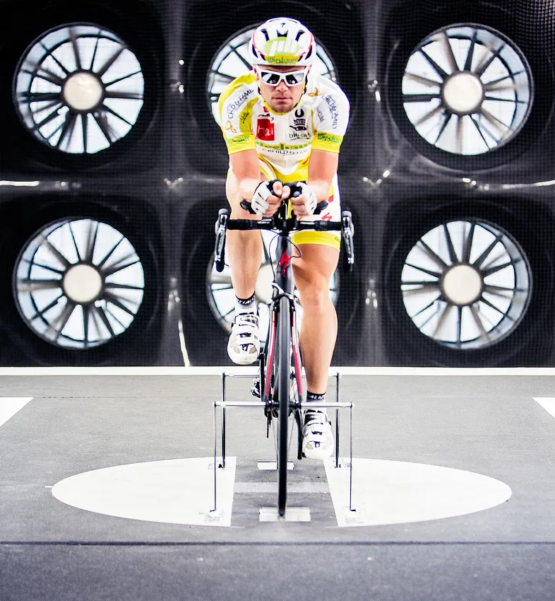 Un cycliste en position aérodynamique sur un vélo d'entraînement dans un tunnel à vent, testant les performances de vêtements de sport sur mesure.