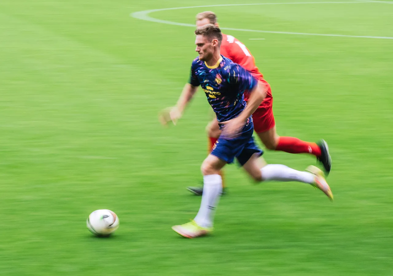 Un joueur de football en maillot coloré sprinte sur l'herbe tandis qu'un autre joueur le suit de près.