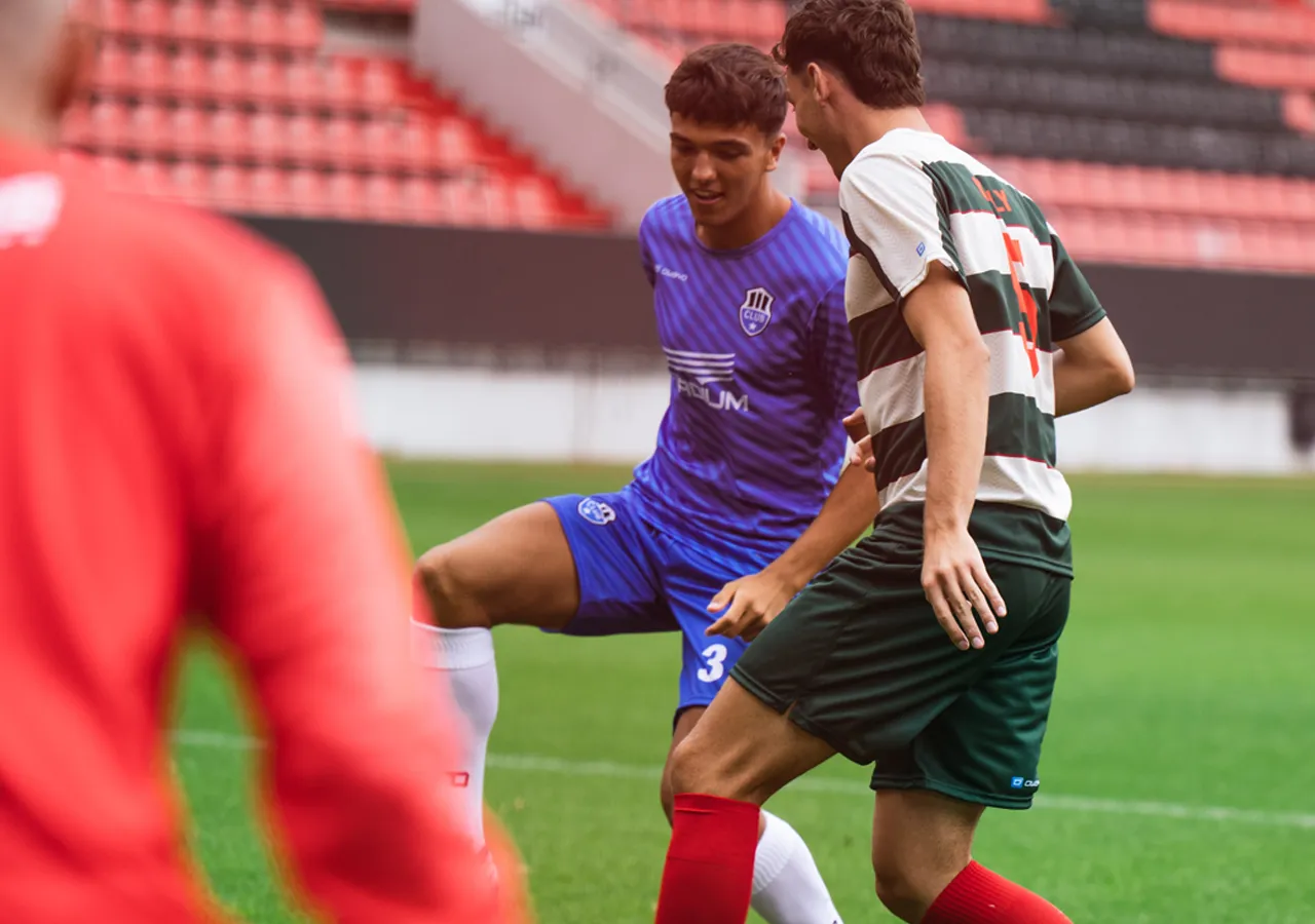 Deux joueurs de football en action sur le terrain, l'un portant un maillot bleu et l'autre un maillot rayé vert et blanc.