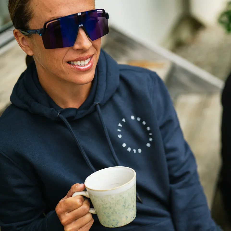 Une personne portant un sweat à capuche bleu foncé avec un logo sourit à la caméra tout en tenant une tasse.
