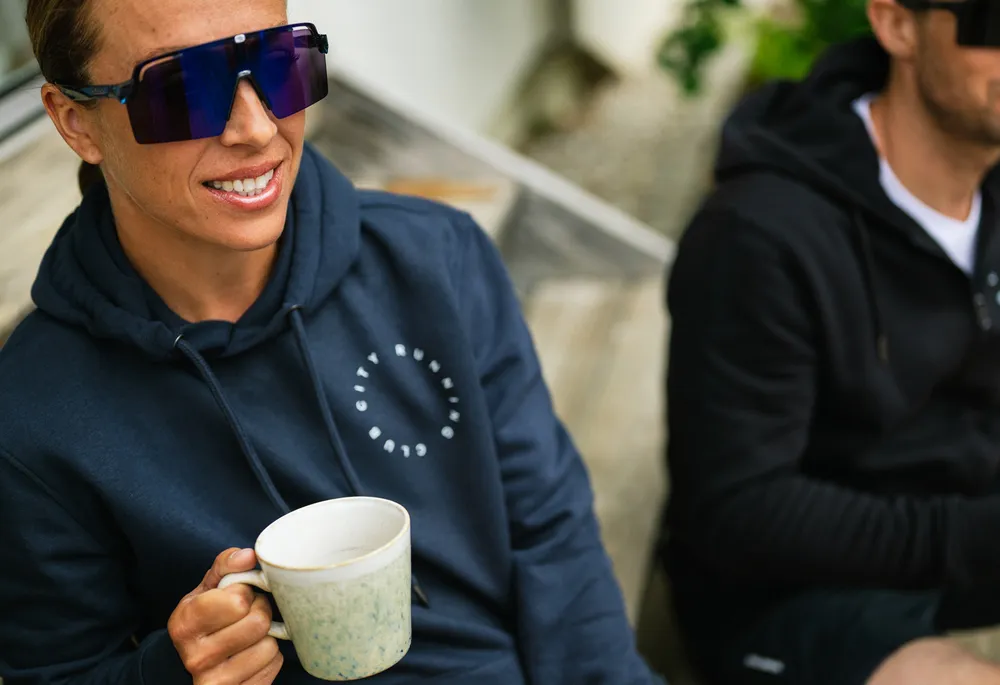 Une personne portant un hoodie bleu avec un logo tient une tasse en s'asseyant sur un banc en bois.