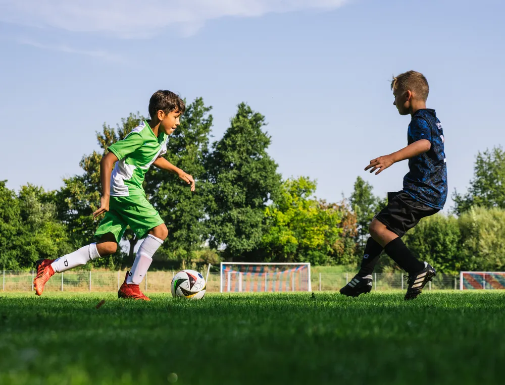Deux enfants jouent au football sur un terrain vert, l'un portant un maillot vert et l'autre un maillot noir, tandis qu'ils poursuivent le ballon.