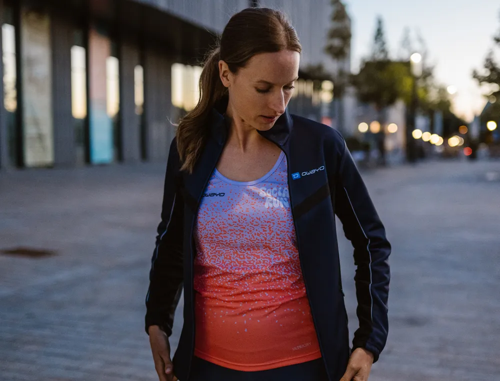 Une femme en tenue de sport, composée d'une veste légère et d'un haut coloré, se tient dans un cadre urbain, regardant pensivement sa tenue.