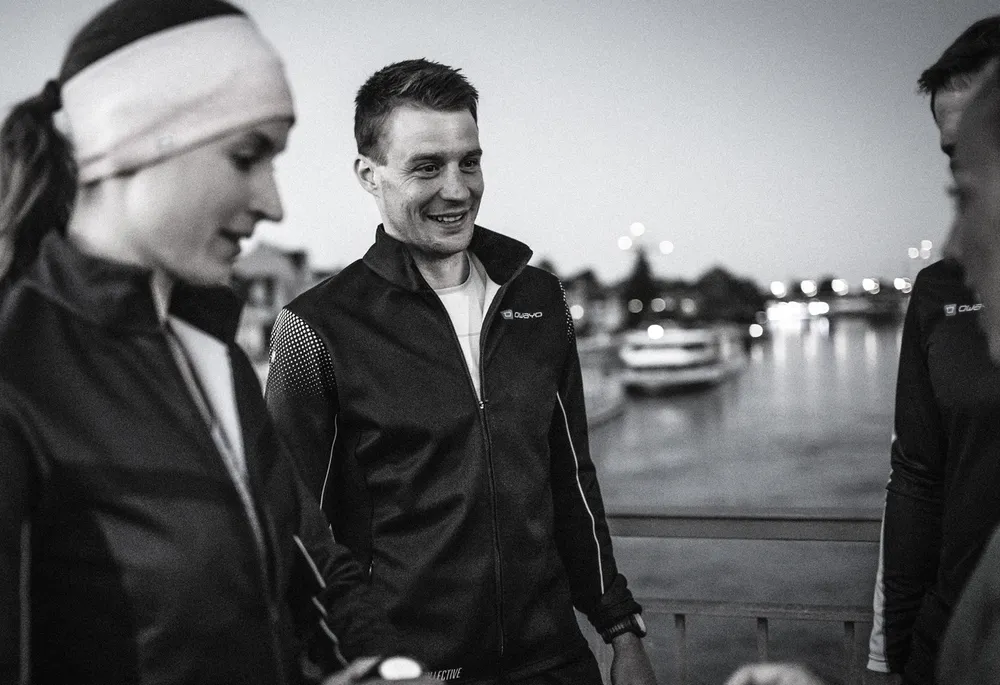 Un groupe d'athlètes portant des vestes de sport sombres se tient au bord de l'eau à l'heure crépusculaire.