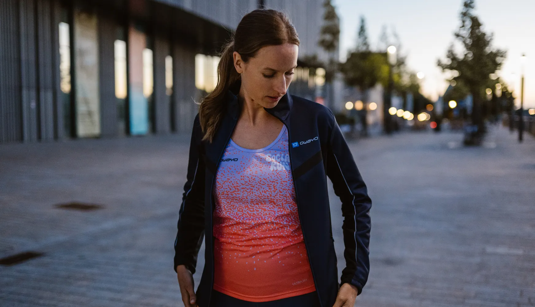 Une femme portant une tenue de sport avec une veste noire et un débardeur coloré et graphique, debout sur un trottoir.