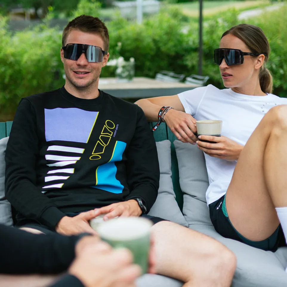 Deux personnes sont assises sur un canapé d'extérieur, l'homme portant un sweat noir avec un design coloré et la femme en t-shirt blanc, tous deux tenant des boissons.