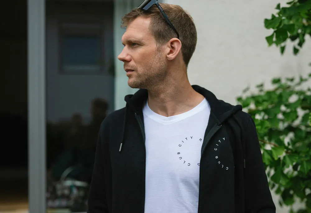 Un homme se tient de côté portant un hoodie noir et un t-shirt blanc avec un imprimé circulaire.