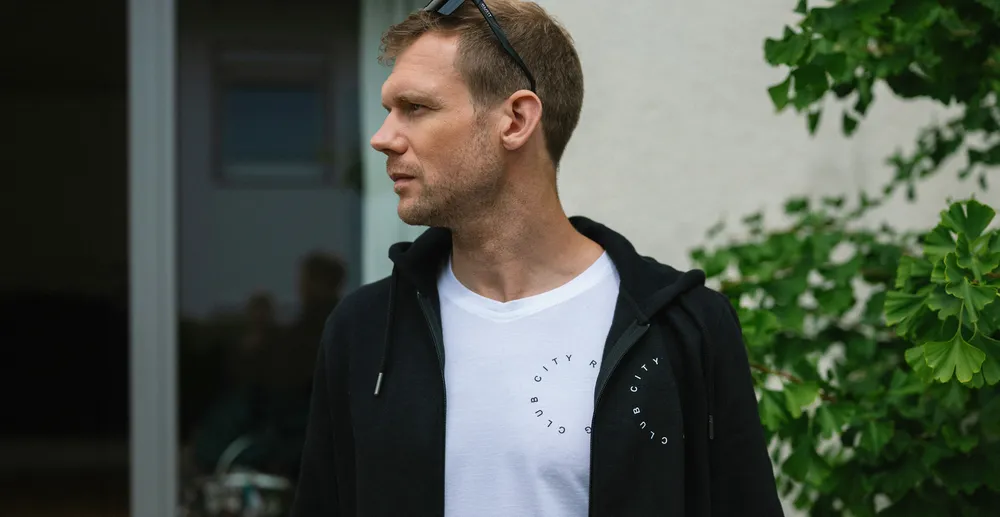 Un homme se tient de côté, portant un hoodie noir sur un T-shirt blanc avec une impression, entouré de plantes.