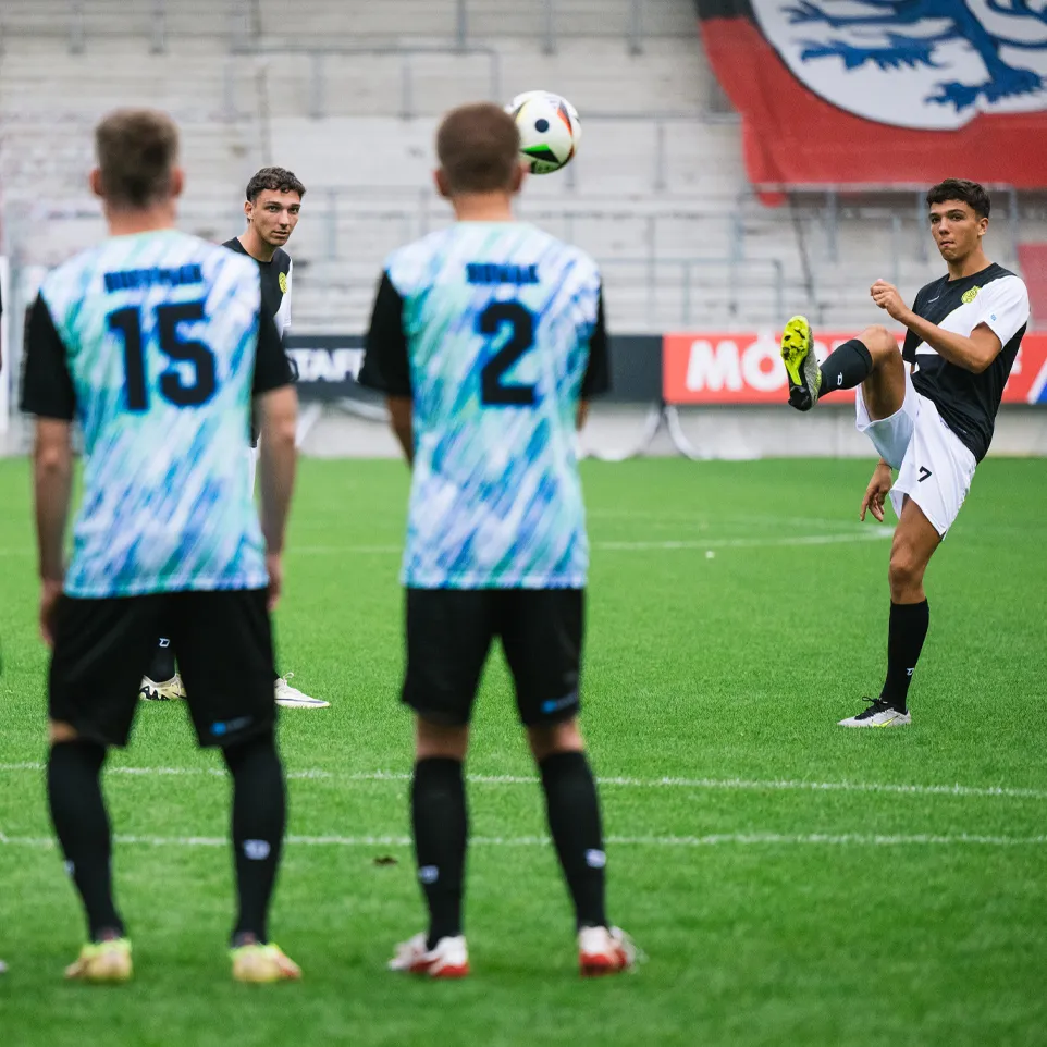 Trois footballeurs en maillots colorés se tiennent sur le terrain et regardent un autre joueur jongler avec un ballon.