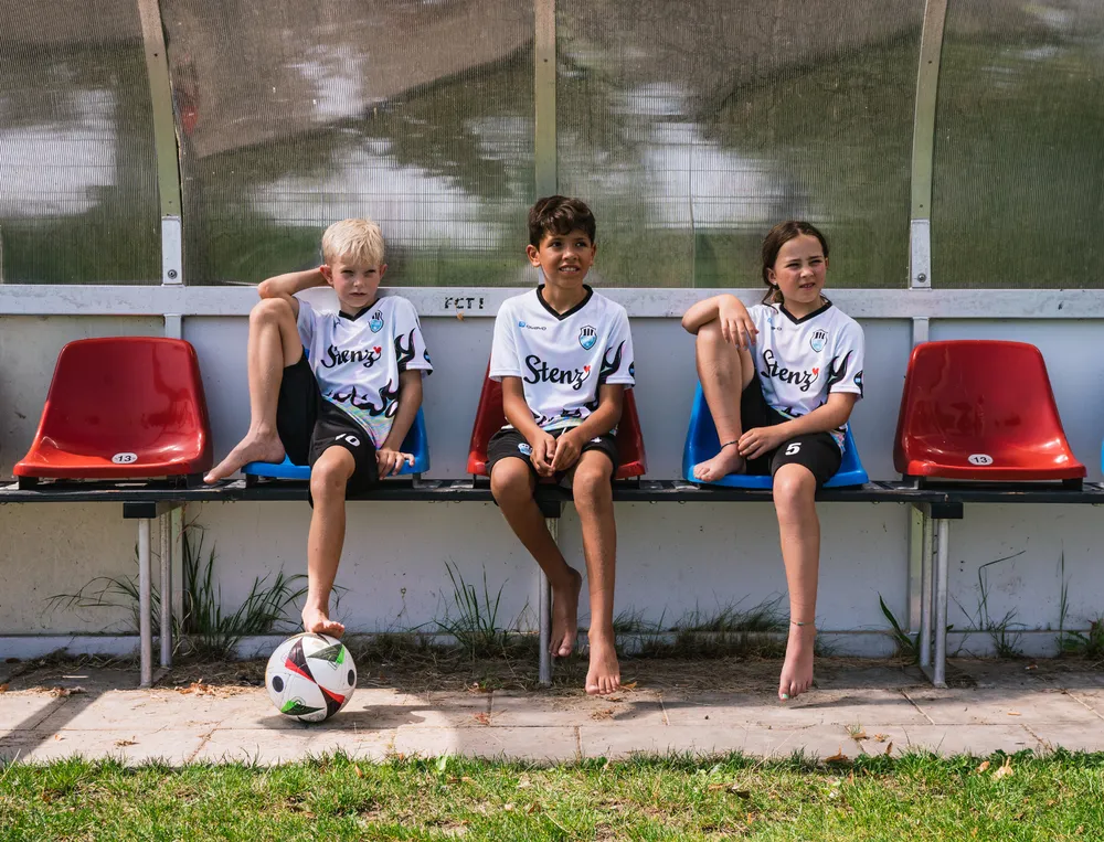Trois enfants sont assis sur un banc au bord d'un terrain de football, portant des maillots de football personnalisés avec un ballon de football devant eux.