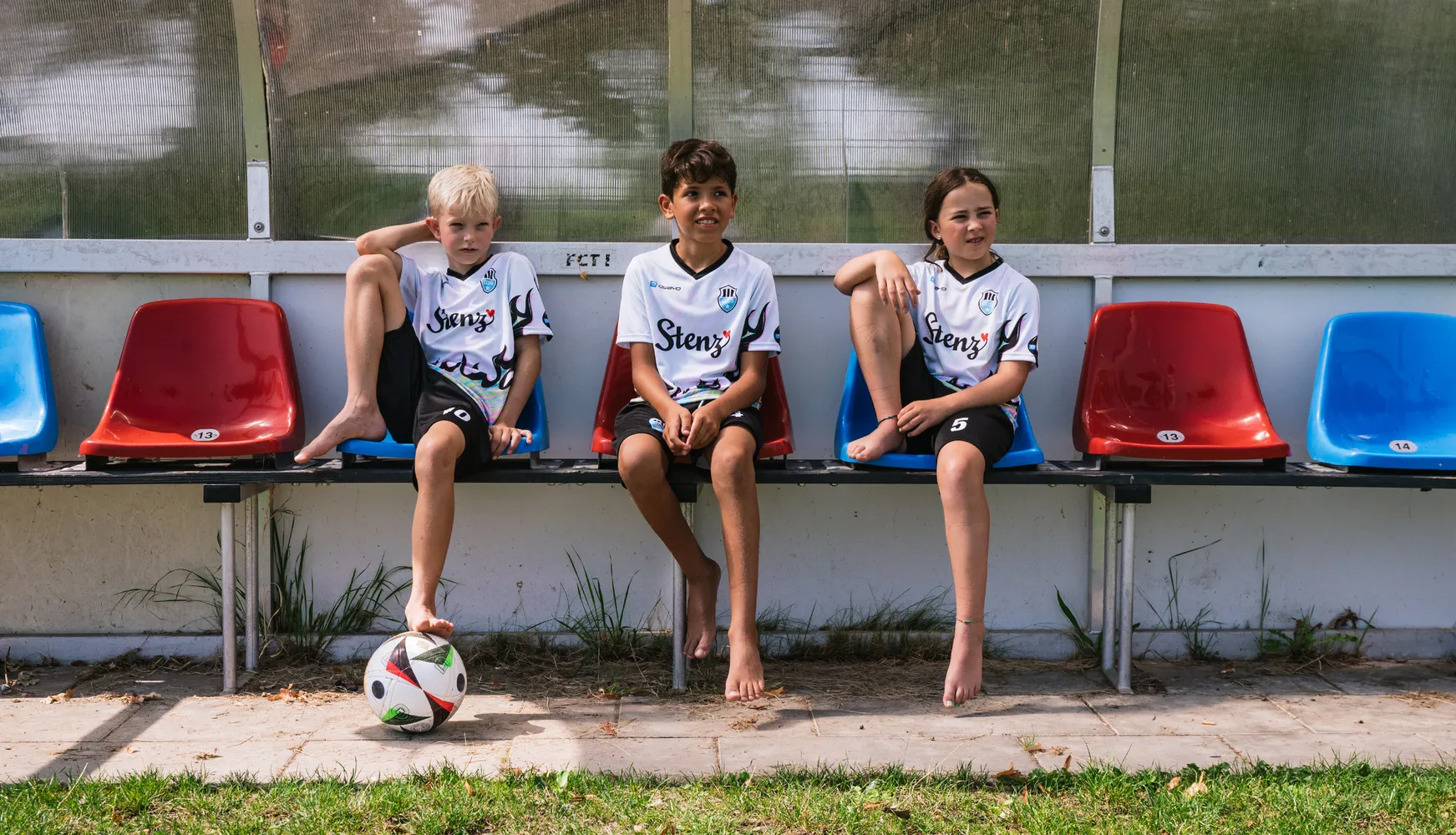 Trois enfants portant des maillots de soccer personnalisés sont assis sur un banc avec un ballon de soccer au sol.