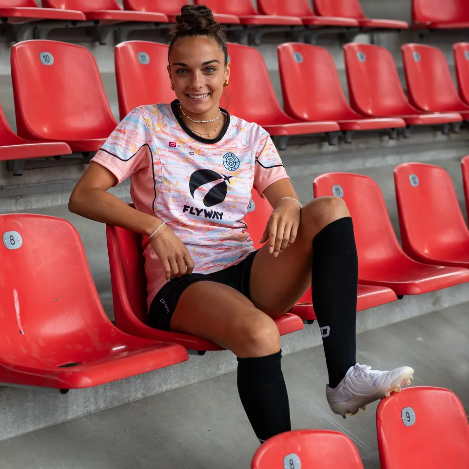 Une femme souriante est assise dans un stade sur un siège rouge, portant un maillot de football coloré et des chaussettes noires.