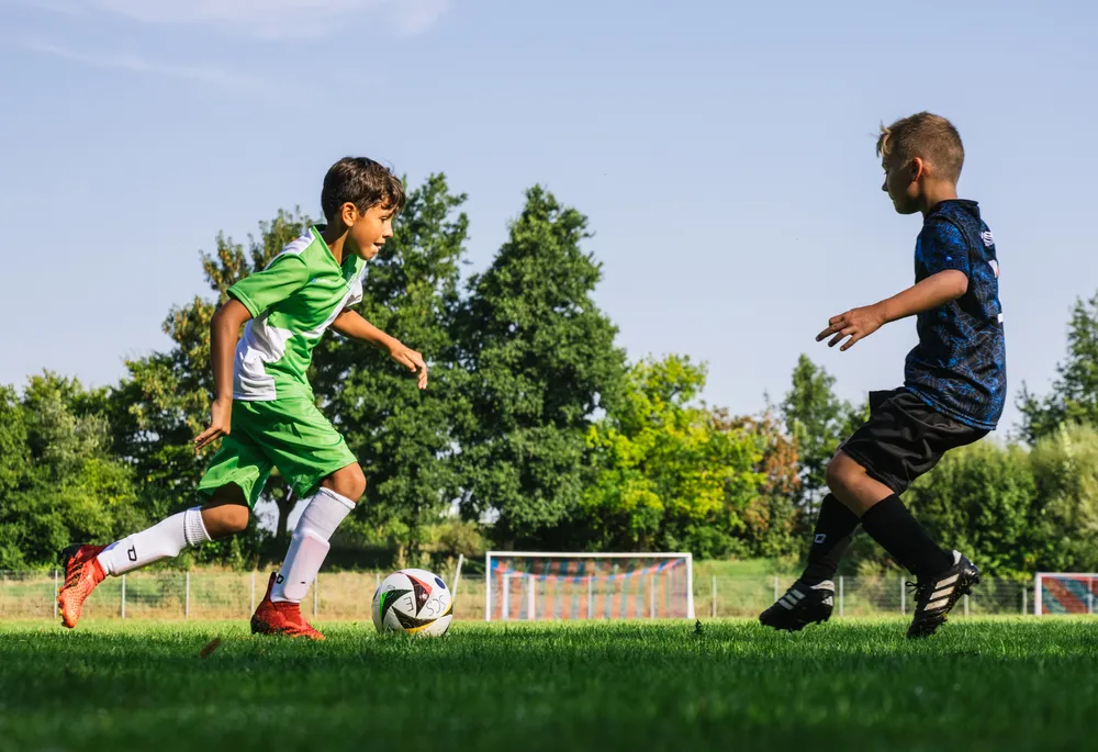 Deux enfants jouent au football sur un terrain vert, l'un portant un maillot vert avec des accents blancs et l'autre un maillot bleu, alors qu'ils poursuivent un ballon.