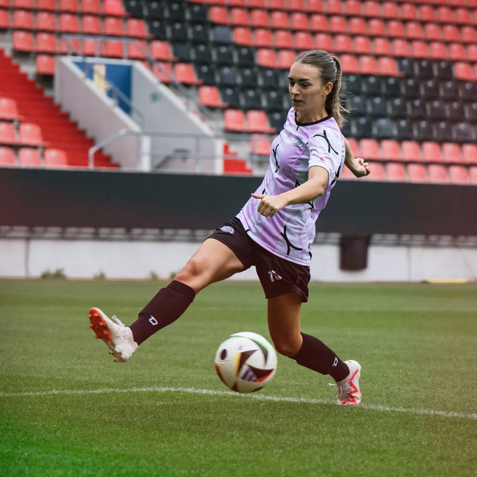 Une joueuse frappe un ballon de football sur un terrain de stade, vêtue d'un maillot violet et de shorts noirs.