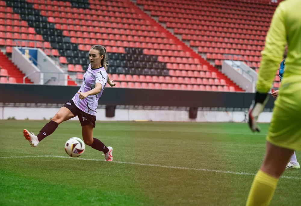 Une footballeuse portant un maillot violet et noir frappe un ballon sur un terrain avec des tribunes vides en arrière-plan.