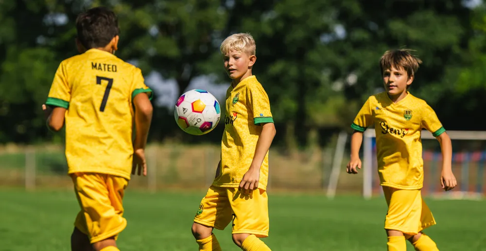 Trois enfants en maillots de football jaunes jouent sur un terrain vert, l'un d'eux contrôlant un ballon de football coloré.