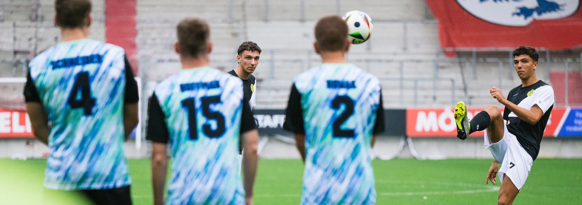 Un groupe de footballeurs en maillots colorés se tient sur le terrain, tandis que l'un d'eux soulève un ballon avec le pied.