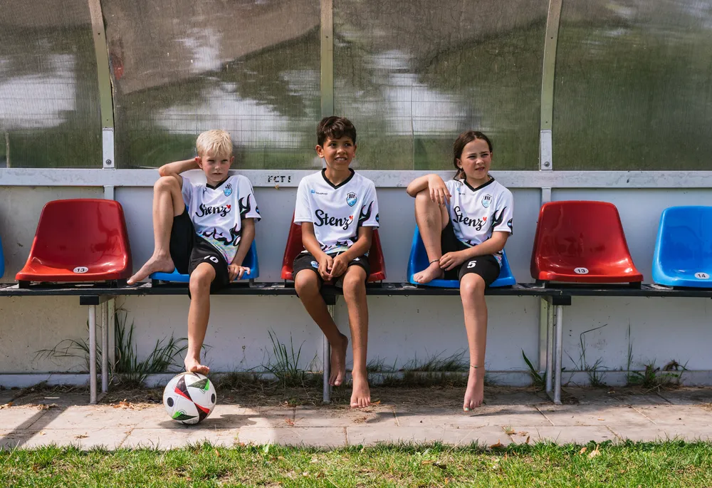 Trois enfants en maillots de football sportifs sont assis sur un banc à côté d'un ballon de football.