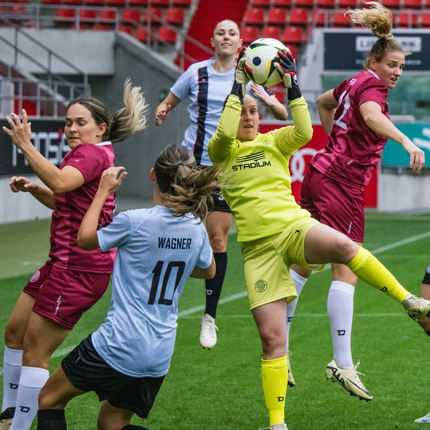 Une gardienne en tenue de sport jaune attrape le ballon pendant que des joueuses en maillots différents s'affrontent sur le terrain de soccer.