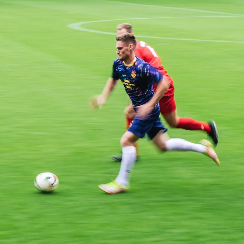 Un joueur de football en maillot coloré sprinte sur le terrain pendant qu'un autre joueur le poursuit.