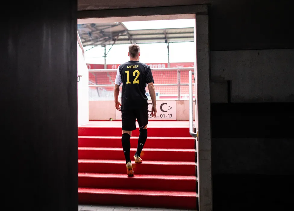 Un footballeur portant un maillot noir avec le numéro 12 monte les escaliers rouges du stade.