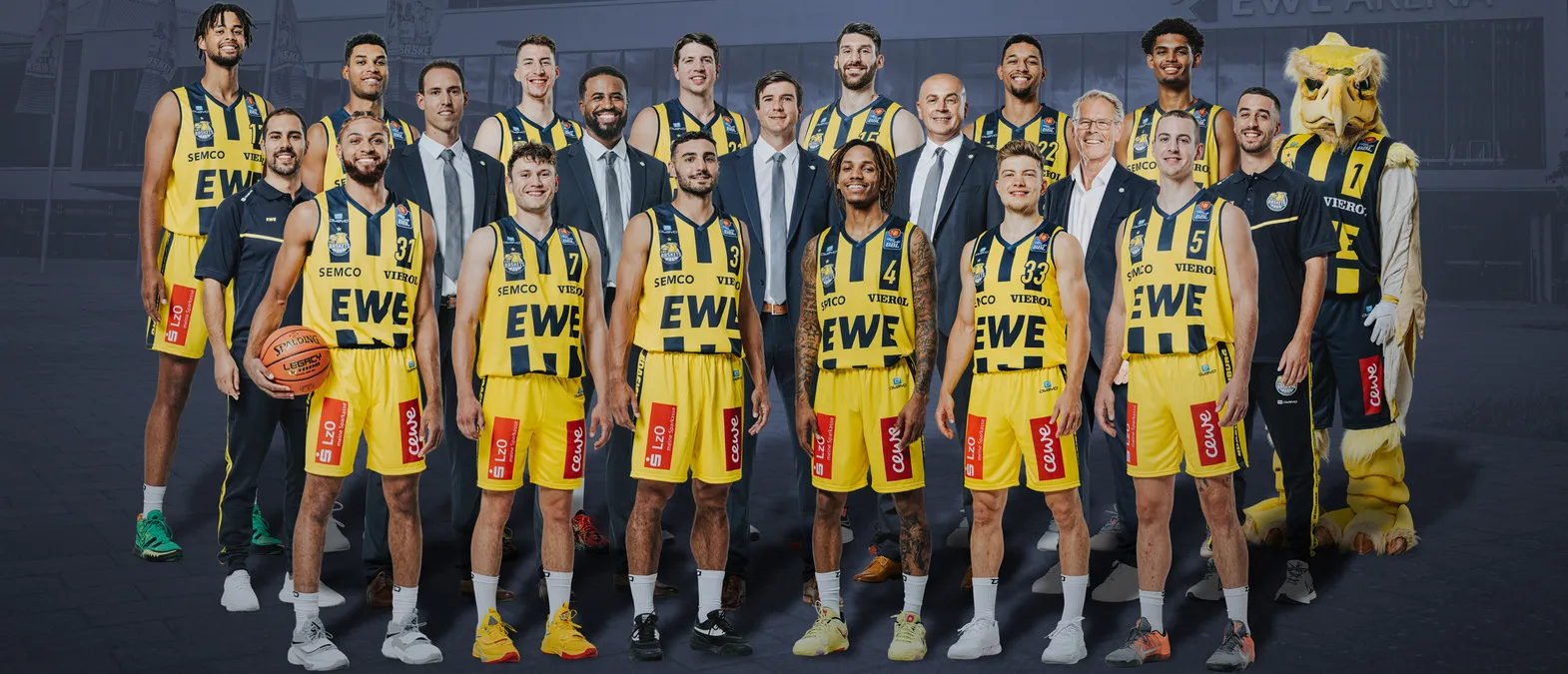 Une photo de groupe d'une équipe de basket-ball portant des maillots jaunes et noirs avec les sponsors 'EWE' et 'SEMC', un mascotte et un ballon de basket, devant un bâtiment.