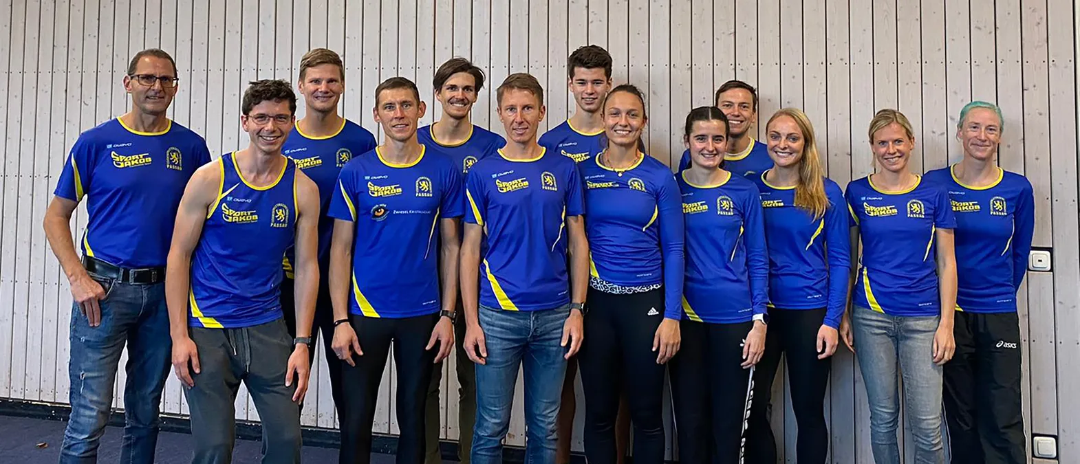 Un groupe d'hommes et de femmes en vêtements de sport bleus et jaunes avec le logo 'Sport Jakob Passau' posent devant un mur en bois clair.