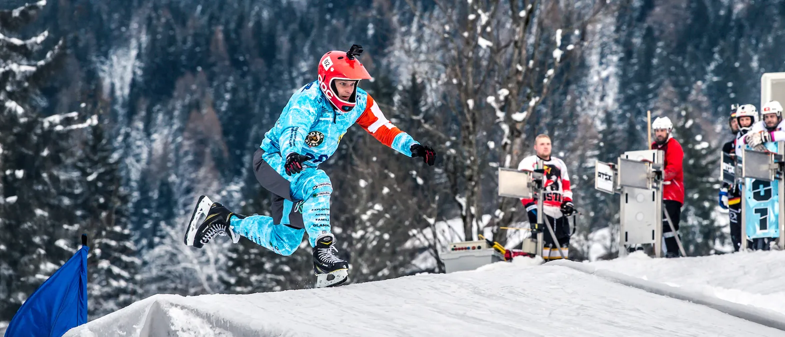 Un joueur de hockey sur glace portant un maillot bleu et blanc personnalisé avec des logos de sponsors et un casque rouge saute par-dessus une rampe de neige sur une patinoire, avec d'autres joueurs et des arbres en arrière-plan.