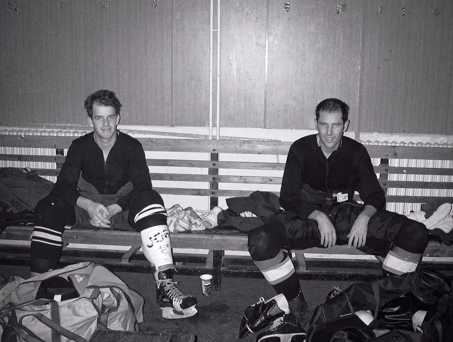 Deux joueurs de hockey sur glace en équipement complet sont assis sur un banc en bois dans un vestiaire, entourés de sacs de sport et de patins.