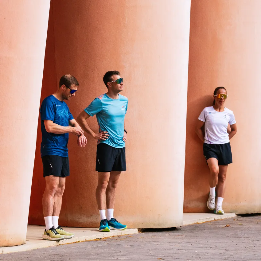 Trois athlètes en vêtements de sport et lunettes de soleil se tiennent décontractés contre des colonnes roses.