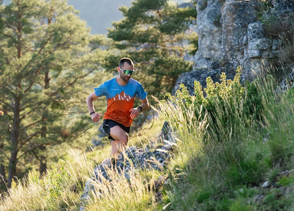 Un coureur en t-shirt coloré et short courant sur un chemin rocheux dans un paysage boisé verdoyant.