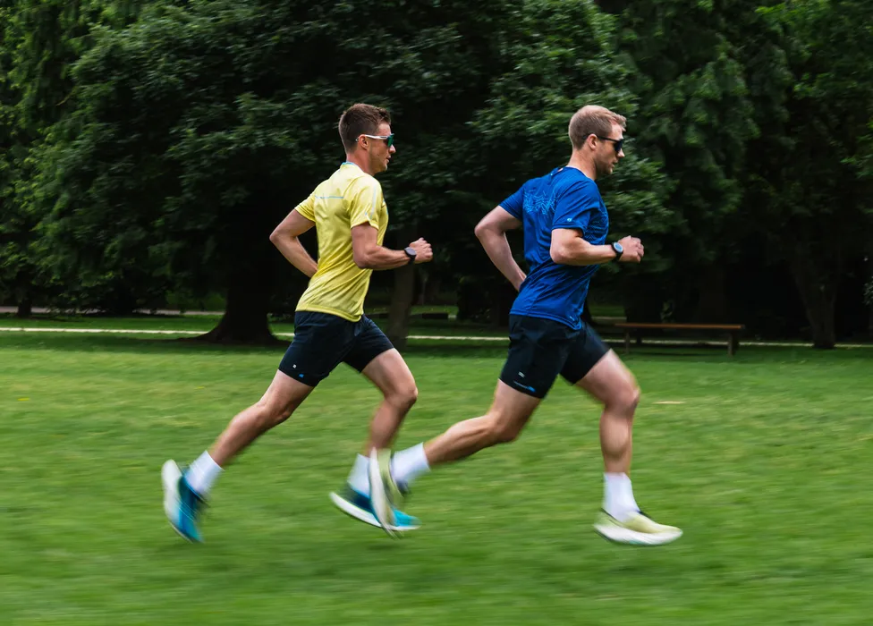 Deux hommes en vêtements de sport courent sur un champ herbeux, l'un portant un t-shirt jaune et l'autre un t-shirt bleu.