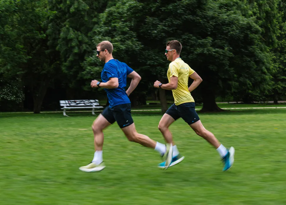 Deux hommes en tenue de sport courent sur un champ vert, l'un portant un t-shirt bleu et l'autre un t-shirt jaune.