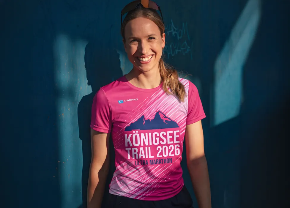 Une femme souriante porte un t-shirt rose avec l'impression 'Königsee Trail 2026 Ultra Marathon' devant un mur bleu.