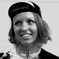 Une femme aux cheveux blonds courts porte une casquette de cyclisme noire avec l'inscription 'VELO LOVE' et un maillot de cyclisme personnalisé avec le nom 'Anja Sturm'.