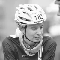 Une cycliste portant un maillot de cyclisme noir avec le logo 'IWC Schaffhausen', un casque blanc avec le numéro 183 et un foulard autour du cou.