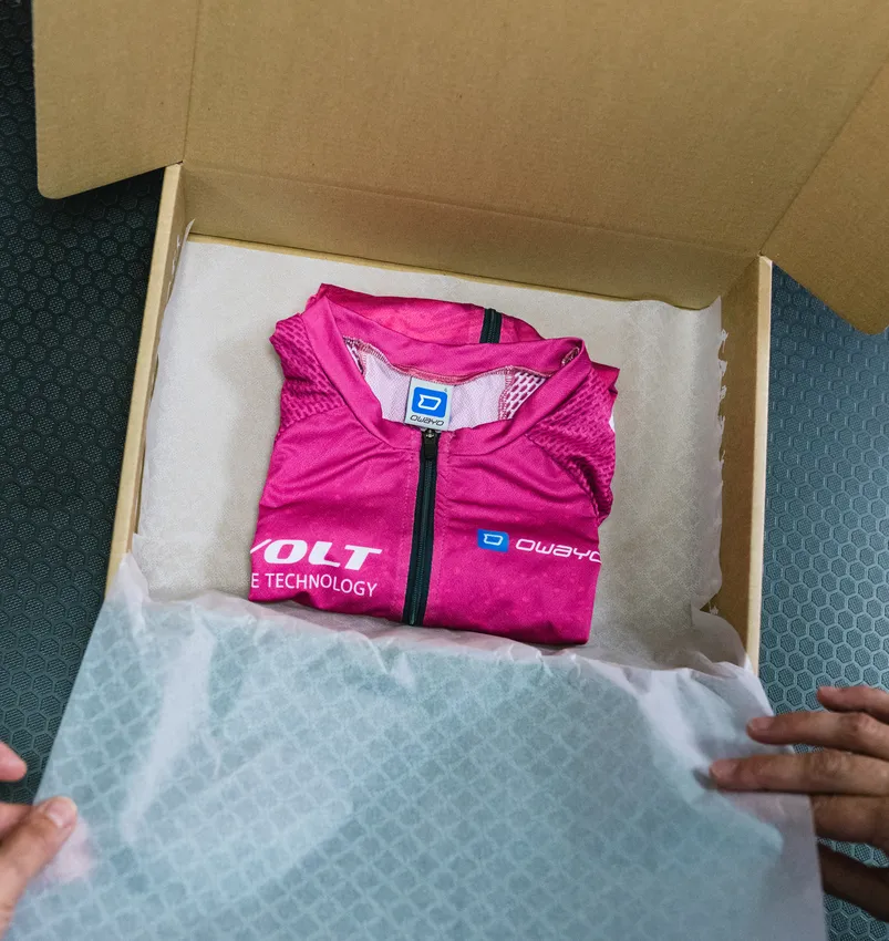 Une tenue de sport personnalisée rose de owayo, pliée, est placée dans une boîte en carton ouverte, sur du papier blanc, tenue par des mains.
