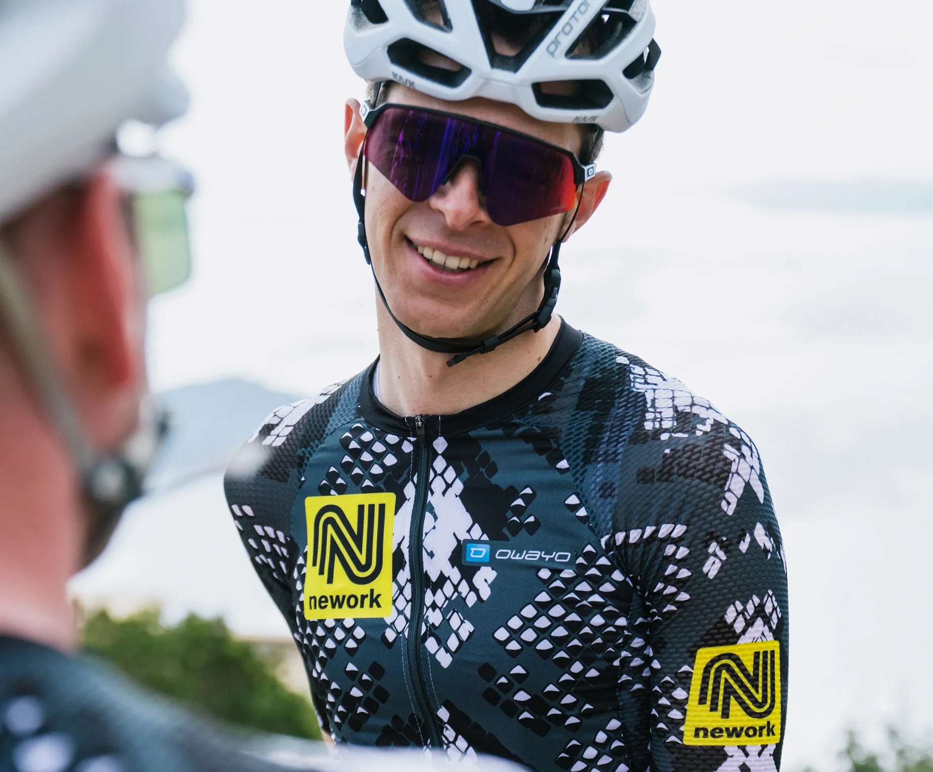 Un cycliste porte un maillot personnalisé à motif noir et blanc avec un logo jaune 'nework' et un logo 'owayo' sur la poitrine.