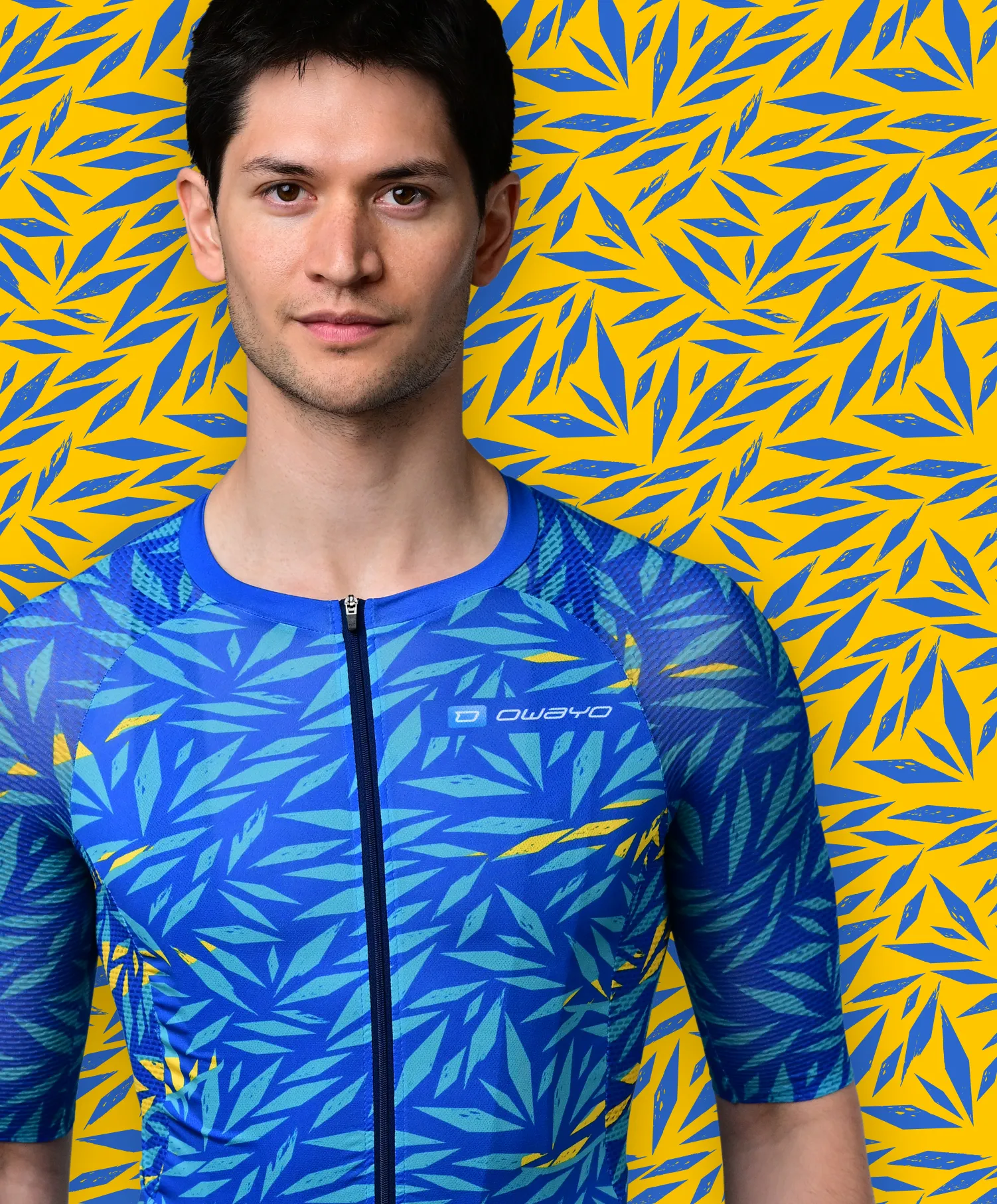 Un homme porte un maillot de sport bleu à manches courtes avec un motif de feuilles bleues et jaunes ressemblant à une plante tropicale, devant un fond jaune avec des motifs de feuilles bleues.