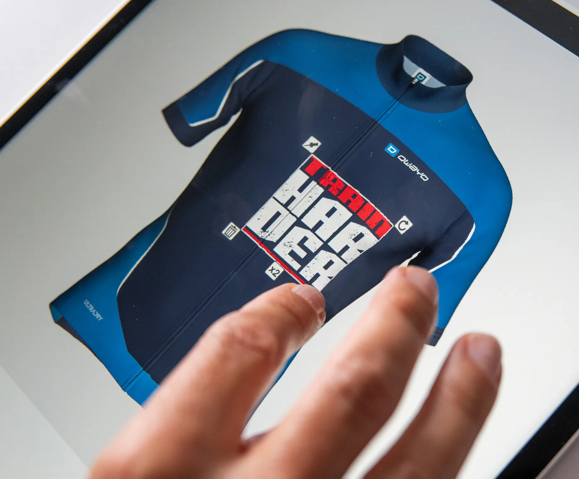 Une main touche un écran affichant un maillot de sport personnalisé bleu foncé et bleu avec le texte 'TRAIN HARDER' et le logo owayo.
