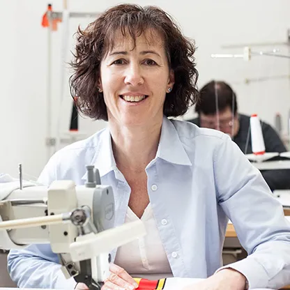 Une femme sourit à la caméra tout en travaillant à une machine à coudre dans un atelier, une autre personne étant visible en arrière-plan également à une machine à coudre.