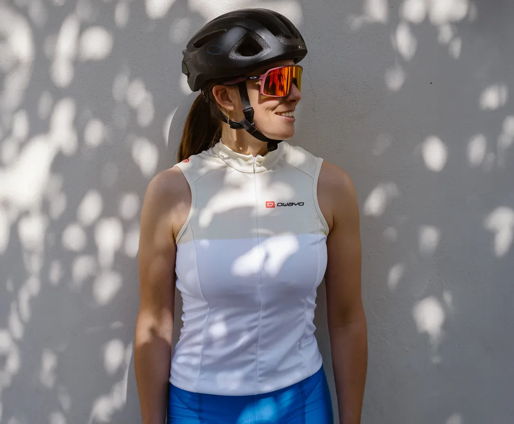 Une femme portant un maillot de cyclisme blanc sans manches avec le logo owayo, un casque de vélo et des lunettes de soleil, appuyée contre un mur avec des ombres de feuilles.