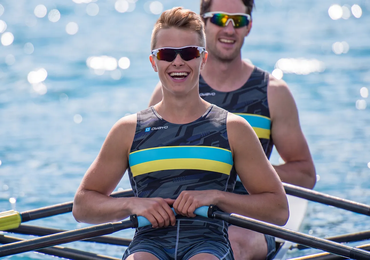 Deux rameurs joyeux en costumes de sport avec des rayures bleues et jaunes assis dans un bateau sur une eau scintillante.